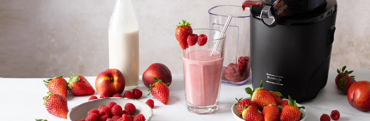 Eye-level panoramic lifestyle shot of a matte black Kuvings juicer next to a tall glass of pink strawberry raspberry smoothie, surrounded by fresh peaches, bowls of berries, and a milk bottle on a white table.