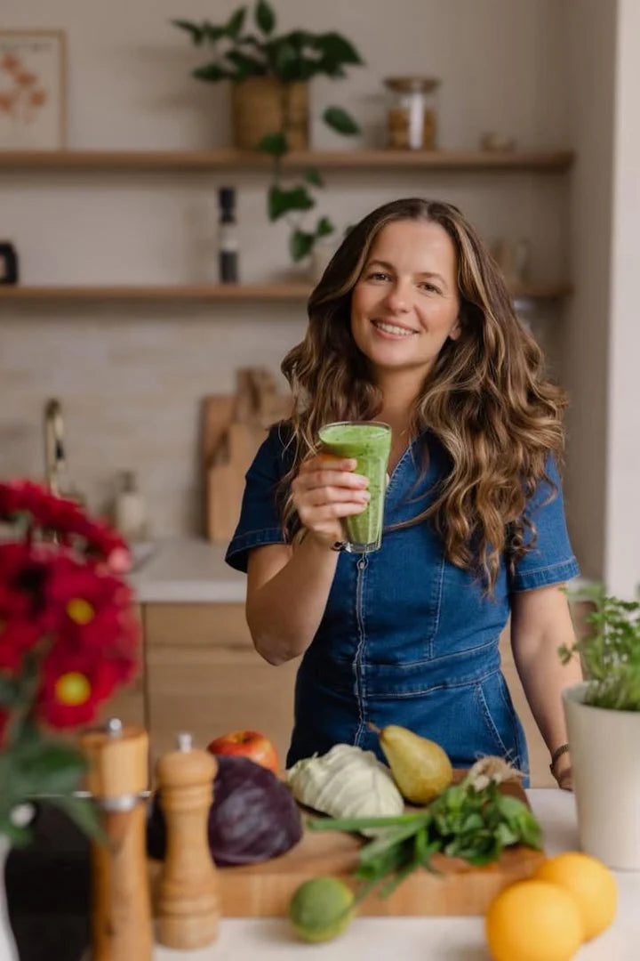 Kuvings green smoothie held by a woman in a kitchen with fresh fruits and vegetables on the counter for healthy homemade smoothie recipes.