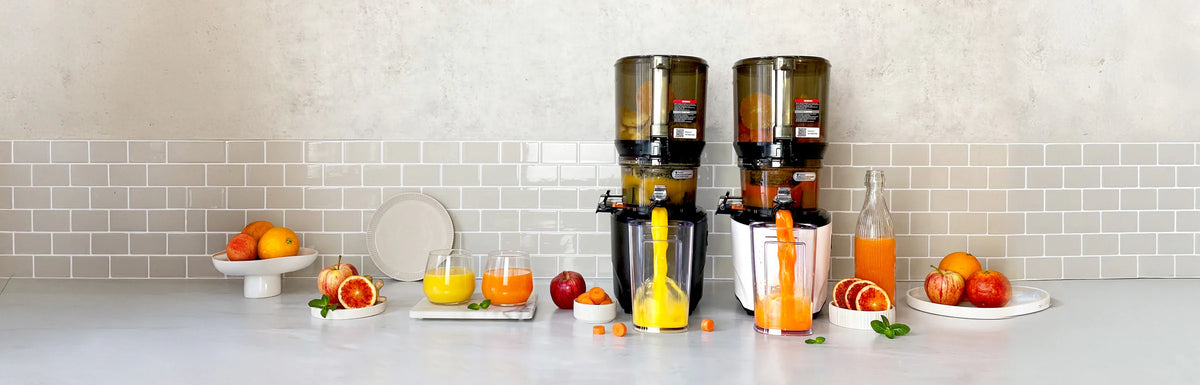 Panoramic banner of a black and a white Kuvings AUTO10S slow juicer side-by-side extracting fresh orange and carrot juice into pitchers on a kitchen counter.