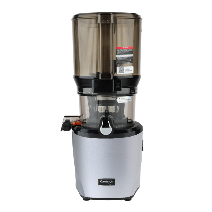 Straight-on studio product shot of a silver Kuvings AUTO10S whole slow juicer with its large transparent grey mega-hopper container attached, showing the integrated handle on the hopper front, isolated against a transparent grid background.