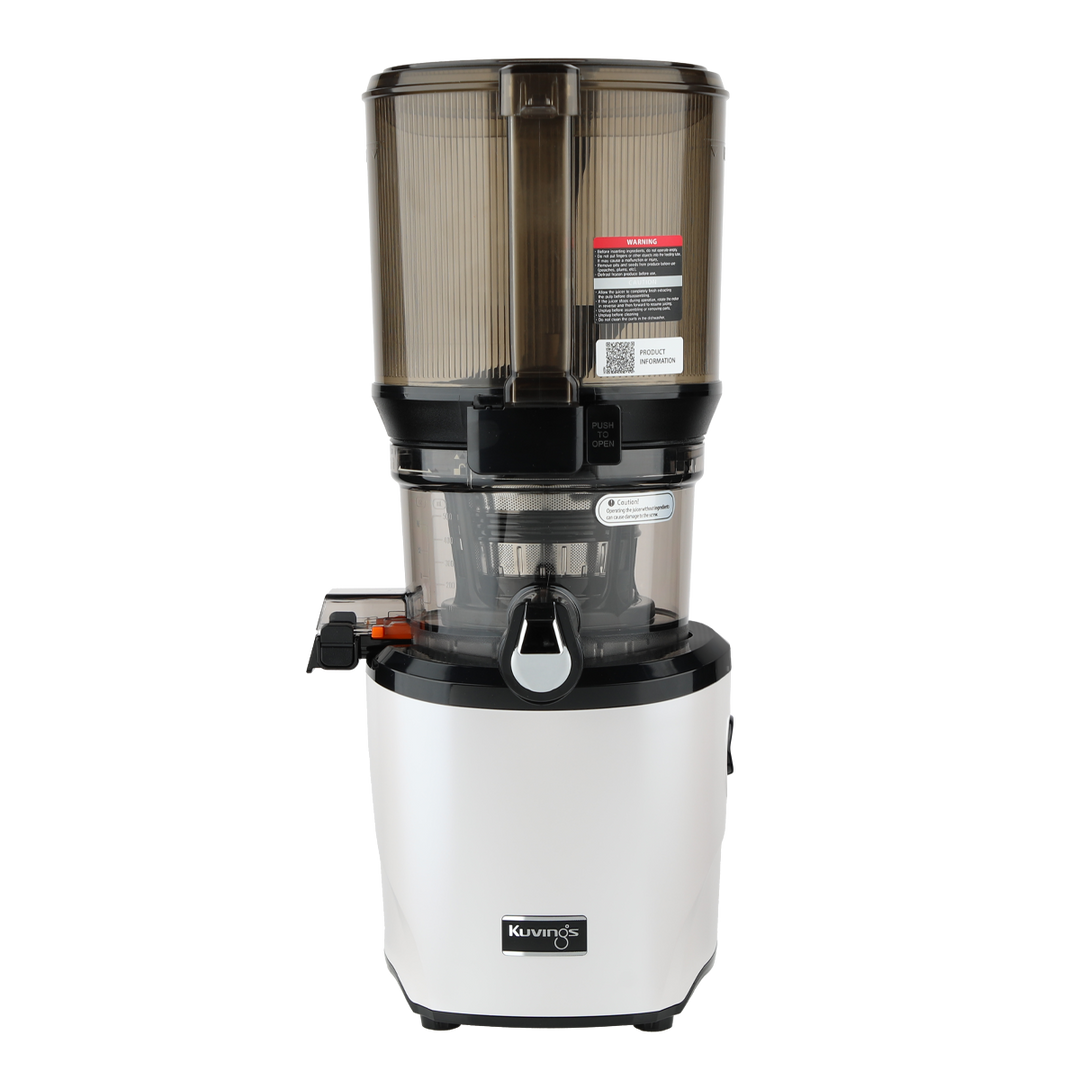 Straight-on studio product shot of a white Kuvings AUTO10S whole slow juicer with its large transparent grey mega-hopper container attached, showing the closed smart cap spout and safety labels, isolated against a transparent grid background.