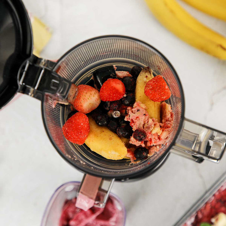 Top-down shot of an open Kuvings AUTO6 slow juicer hopper filled with whole strawberries, bananas, and blueberries for making a fruit sorbet.
