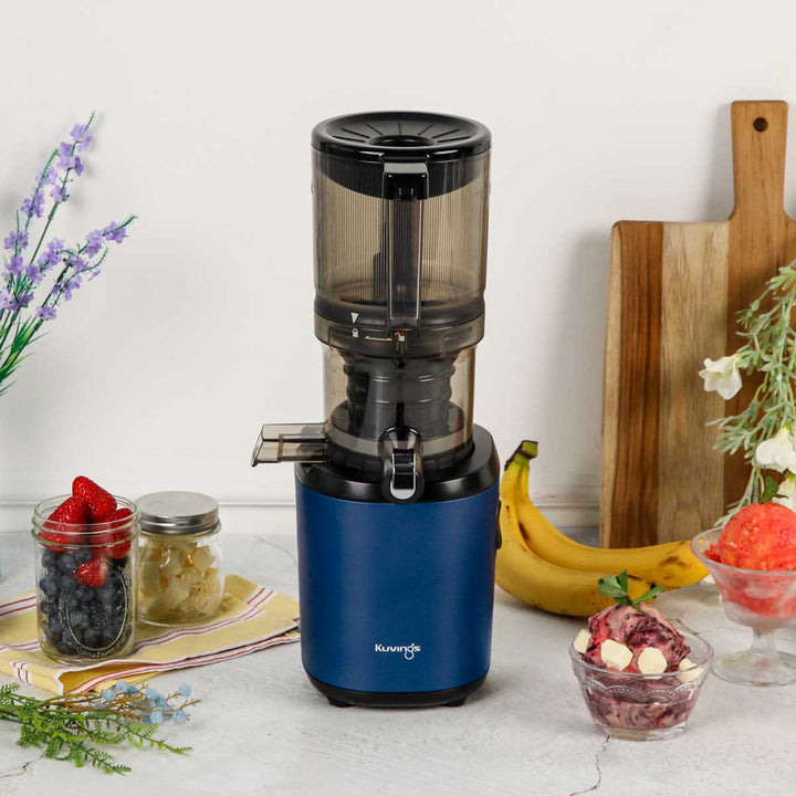 Lifestyle kitchen shot of a navy blue Kuvings AUTO6 slow juicer surrounded by fresh bananas, a jar of berries, and bowls of homemade fruit sorbet.