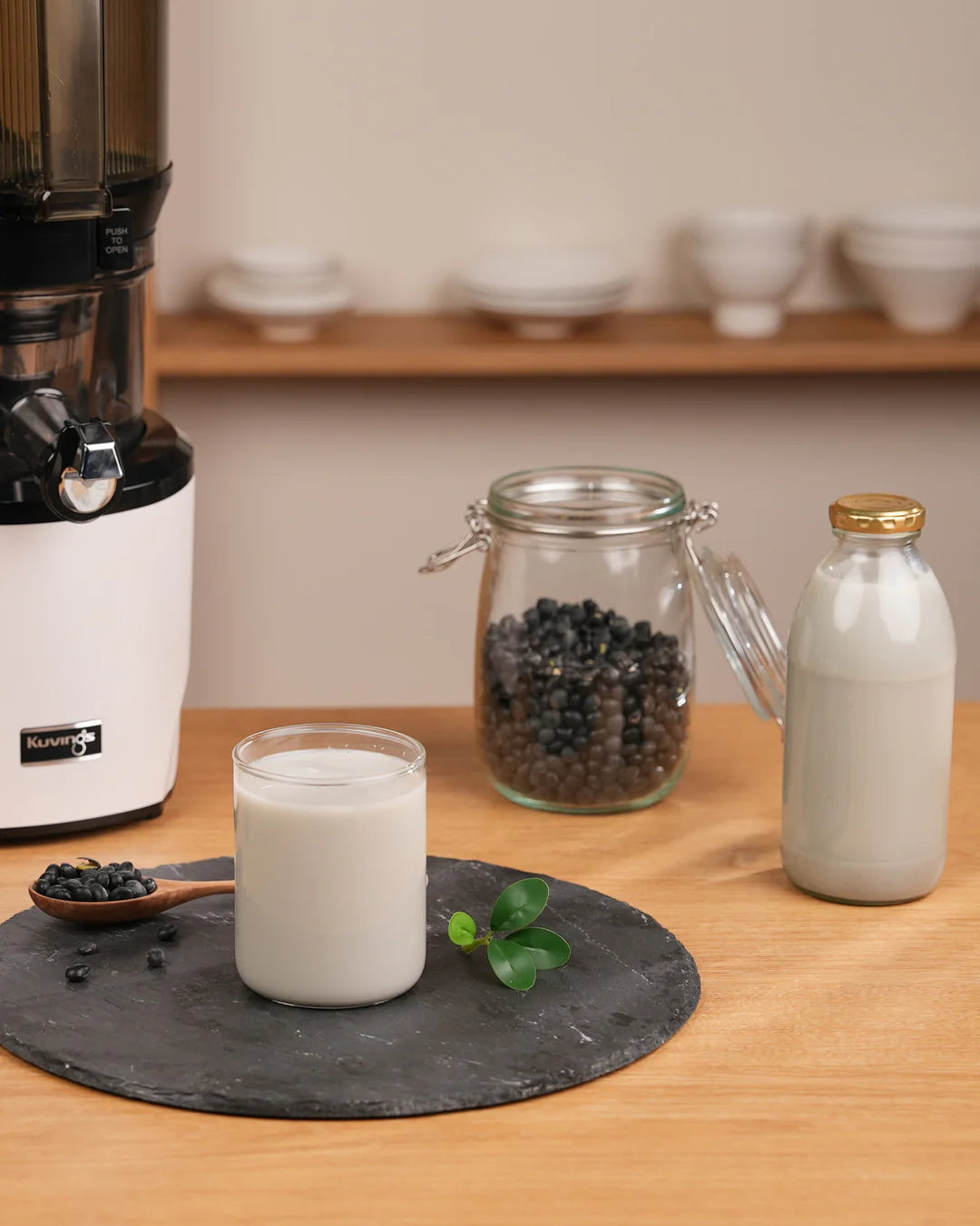 Kuvings AUTO10 slow juicer with black soy milk, soybeans and bottled plant-based milk on a kitchen counter.