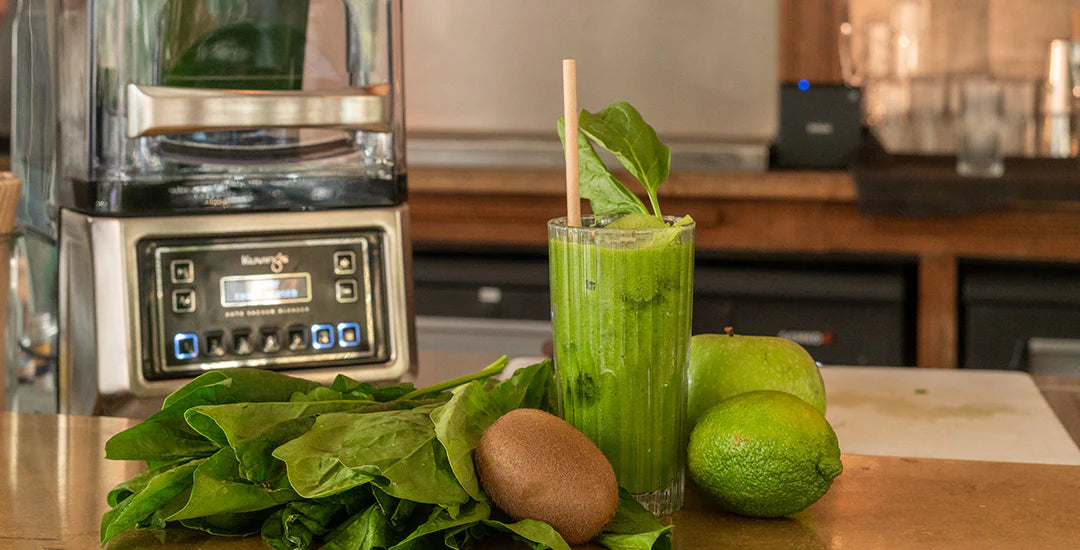 Eye-level wide shot of a fresh green smoothie in a tall glass surrounded by spinach leaves, kiwi, lime, and a green apple on a counter, with a Kuvings CB1000 commercial vacuum blender in the background.