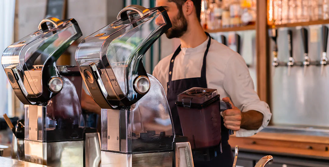 Widok z poziomu oczu, średni kadr, dwa profesjonalne blendery próżniowe Kuvings CB1000 z otwartymi osłonami wyciszającymi na ladzie barowej, z męskim baristą w fartuchu trzymającym dzbanek blendera w tle.