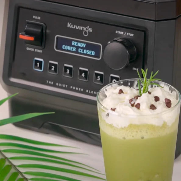 Kosi pogled izbliza na svježi smoothie od zelenog čaja preliven šlagom i komadićima čokolade, postavljen ispred Kuvings CB850 blendera koji na digitalnom zaslonu prikazuje 'READY COVER CLOSED'.