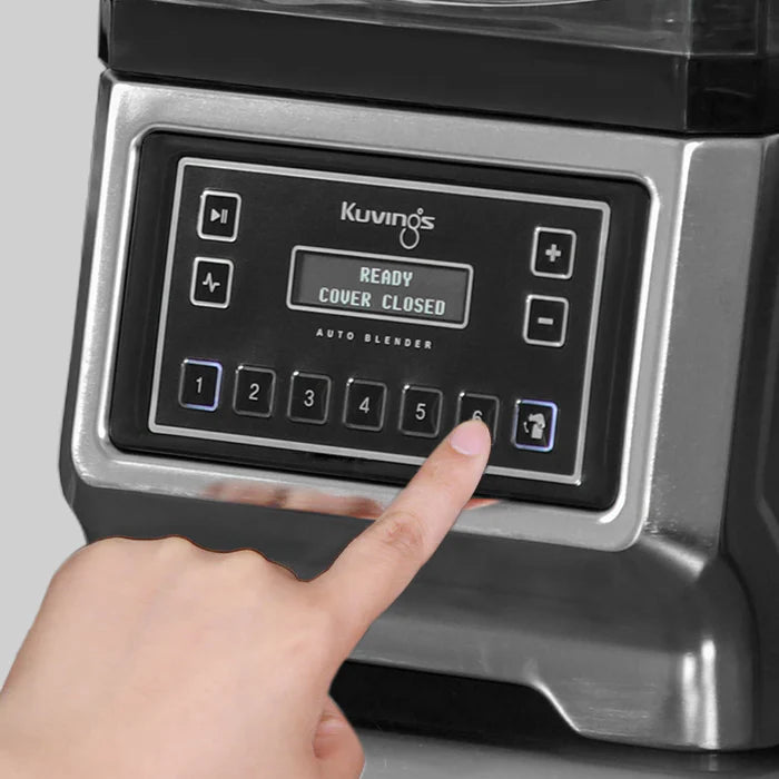 Widok pod kątem, z bliska, palec naciskający przycisk szósty na srebrnej metalowej konsoli sterowania automatycznego blendera Kuvings CB980, z wyświetlaczem cyfrowym pokazującym 'READY COVER CLOSED'.