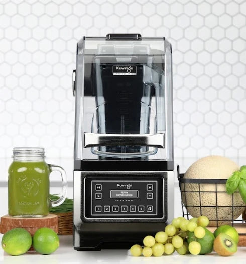 Widok z przodu, na poziomie oczu, profesjonalny automatyczny blender Kuvings CB980 z zamkniętą przezroczystą obudową wyciszającą, stojący na blacie kuchennym obok słoika typu mason z zielonym sokiem i świeżymi owocami na tle białej mozaiki heksagonalnej.