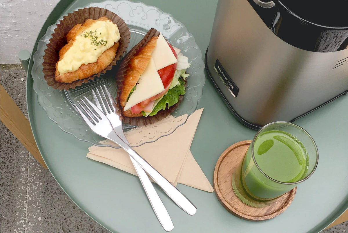 Oben-von-oben-Ansicht einer Frühstücksszene auf einem salbeigrünen runden Tisch, mit einem klaren Glas frischem grünen Saft auf einem Holzuntersetzer neben dem silbernen Edelstahl-Sockel eines Kuvings Chef CS700 gewerblichen Juicers, gepaart mit einem Glasteller, auf dem zwei gefüllte Croissants und zwei Metallgabeln liegen.