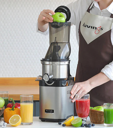 Mittlere Aufnahme auf Augenhöhe einer Person, die eine braune Kuvings-Schürze trägt und einen ganzen grünen Apfel in den oberen Klappdeckel eines Edelstahl-Kuvings Chef CS700 gewerblichen Slow Juicers einlegt, umgeben von Gläsern mit verschiedenen Fruchtsäften und frischen Produkten auf einer Arbeitsplatte.