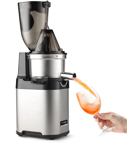 Produktaufnahme eines Edelstahl-Kuvings Chef CS700 Gewerbe-Slow-Juicers von dreiviertel Ansicht mit angebrachtem Deckel des breiten Klapptor-Einfüllschachts, der zeigt, wie eine Hand ein Weinglas kippt, um frischen rosa Grapefruit-Saft aufzufangen, der aus dem offenen Metallauslauf auf weißem Hintergrund fließt.