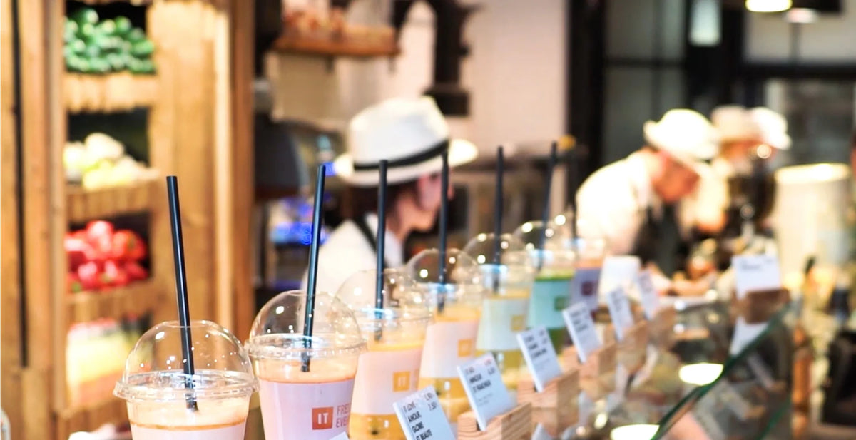 Ujęcie na poziomie oczu z małą głębią ostrości, przedstawiające ruchliwy blat baru z sokami, pokazujące rząd świeżych soków i smoothie w przezroczystych plastikowych kubkach ze słomkami ustawionych na pierwszym planie, podczas gdy pracownicy w pasujących białych czapkach pracują nieostrzono w tle.