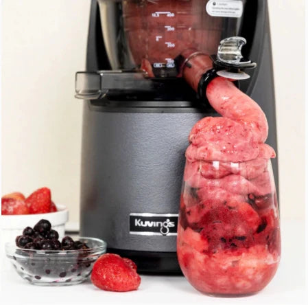 Close-up lifestyle shot of a dark gunmetal grey Kuvings EVO820 slow juicer dispensing a thick, textured frozen red strawberry and berry sorbet directly into a clear glass, with small bowls of fresh strawberries and blueberries resting on the white table surface alongside it.