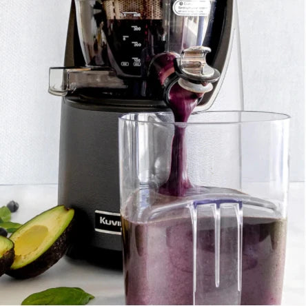 Close-up lifestyle shot of a dark grey Kuvings EVO820 slow juicer pouring a thick purple berry smoothie through its opened smart cap into a clear pitcher, with a halved fresh avocado resting on the table in the foreground.