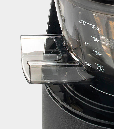 Side close-up studio product shot highlighting the reinforced transparent dark grey pulp outlet chute on the juicing bowl of a Kuvings EVO820 whole slow juicer, showing the printed measurement markings in ounces and milliliters on the side of the drum against a light grey background.