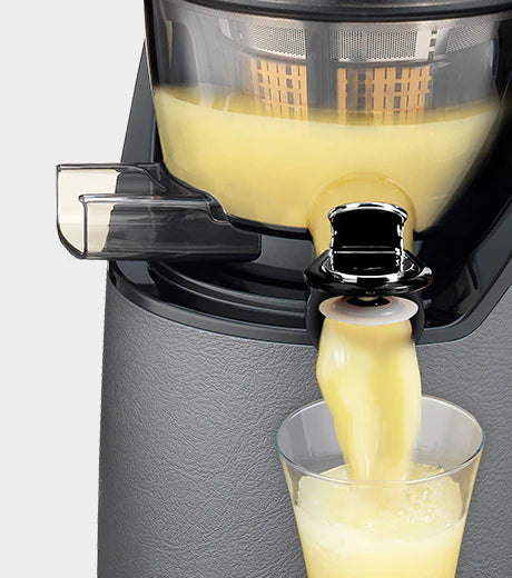 Close-up studio product shot of a gunmetal grey Kuvings EVO820 whole slow juicer with its black chrome smart cap opened, pouring a smooth, bright yellow citrus juice from the transparent juicing bowl into a clear glass against a light grey background.