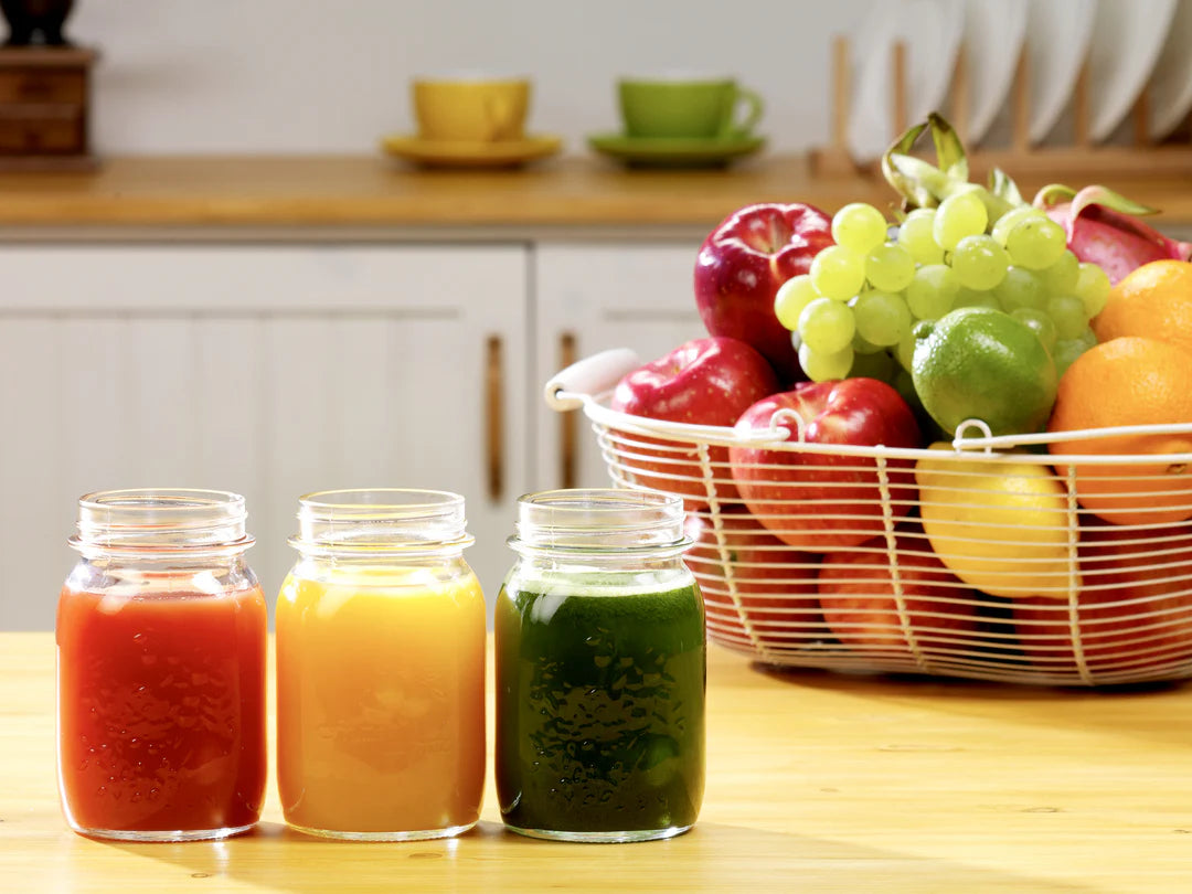 Three jars of juice on a table with a basket of fruits in the background.