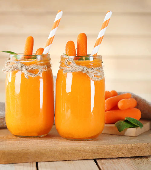 Two jars of carrot juice with straws and carrots on a wooden surface