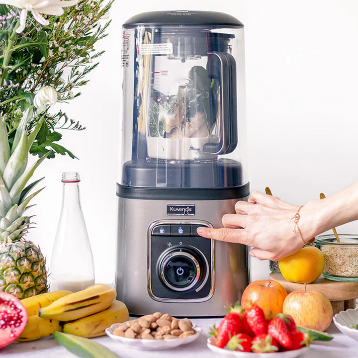 Straight-on, eye-level shot of a hand pressing a preset button on the silver control panel of a Kuvings SV-500 vacuum blender, surrounded by an abundant display of fresh fruits including strawberries, apples, bananas, and a pineapple.