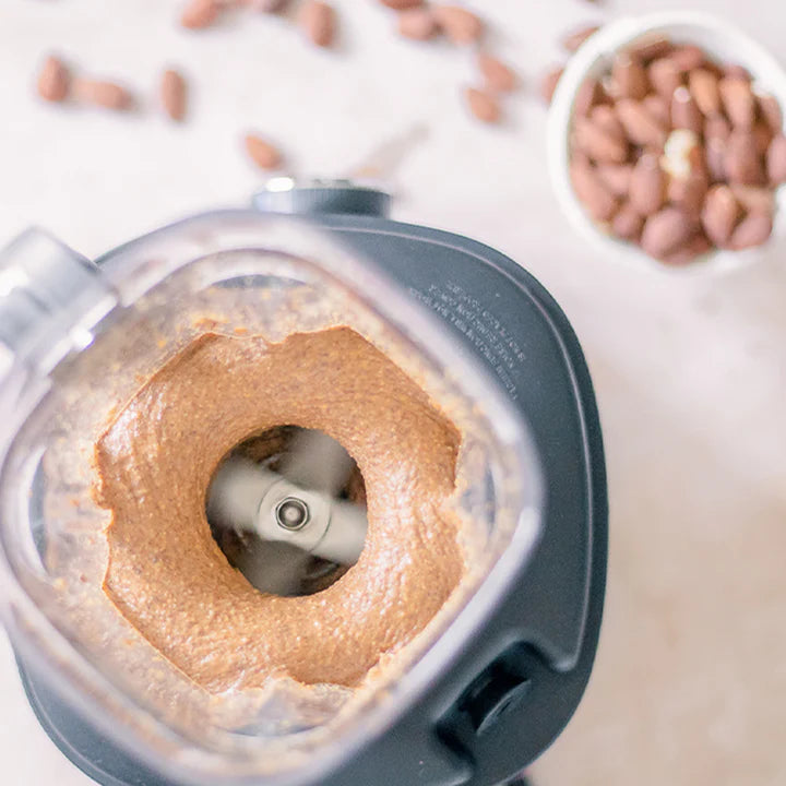 High-angle, overhead view looking down into the open jar of a Kuvings SV-500 vacuum blender as it processes thick, creamy almond butter around its spinning blades, with whole almonds scattered on the light countertop in the background.