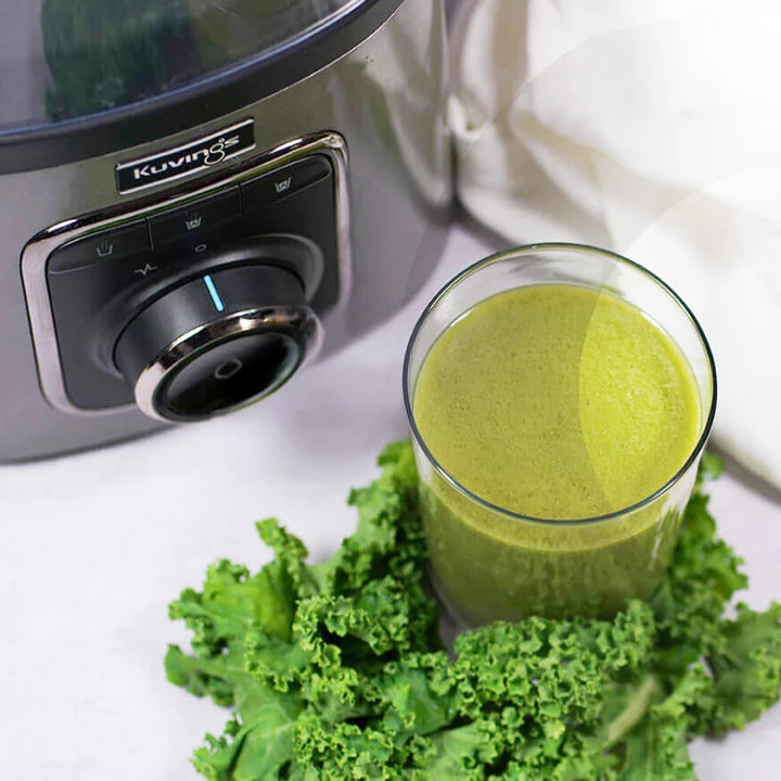 High-angle, close-up view of the silver motor base and control dial of a Kuvings SV-500ME vacuum blender next to a clear glass filled with a vibrant green smoothie resting on a bed of fresh curly kale leaves.