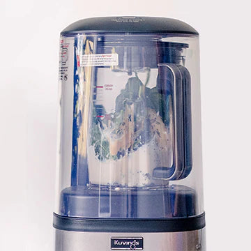 Straight-on, eye-level close-up view of the upper section of a Kuvings SV-500S vacuum blender enclosed within its clear sound shield, showing green leafy vegetables and milk inside the blending jar against a light background.
