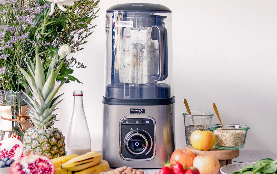 Straight-on, eye-level shot of the Kuvings SV-500S vacuum blender on a kitchen counter blending a creamy mixture, surrounded by an abundant display of fresh fruits including a pineapple, bananas, sliced pomegranates, and apples alongside jars of seeds and grains.