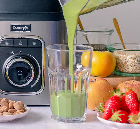Close-up, eye-level shot of a vibrant green smoothie being poured from a Kuvings SV-500S vacuum blender jar into a tall clear glass, positioned next to the blender's silver control panel with fresh strawberries, apples, and almonds on the counter.