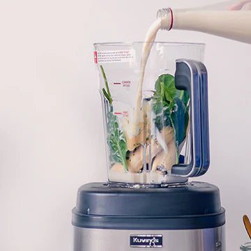 Straight-on, eye-level view of a Kuvings SV-500S vacuum blender jar as plant milk is poured from a glass bottle over fresh green leafy vegetables and fruit pieces inside the container against a light background.