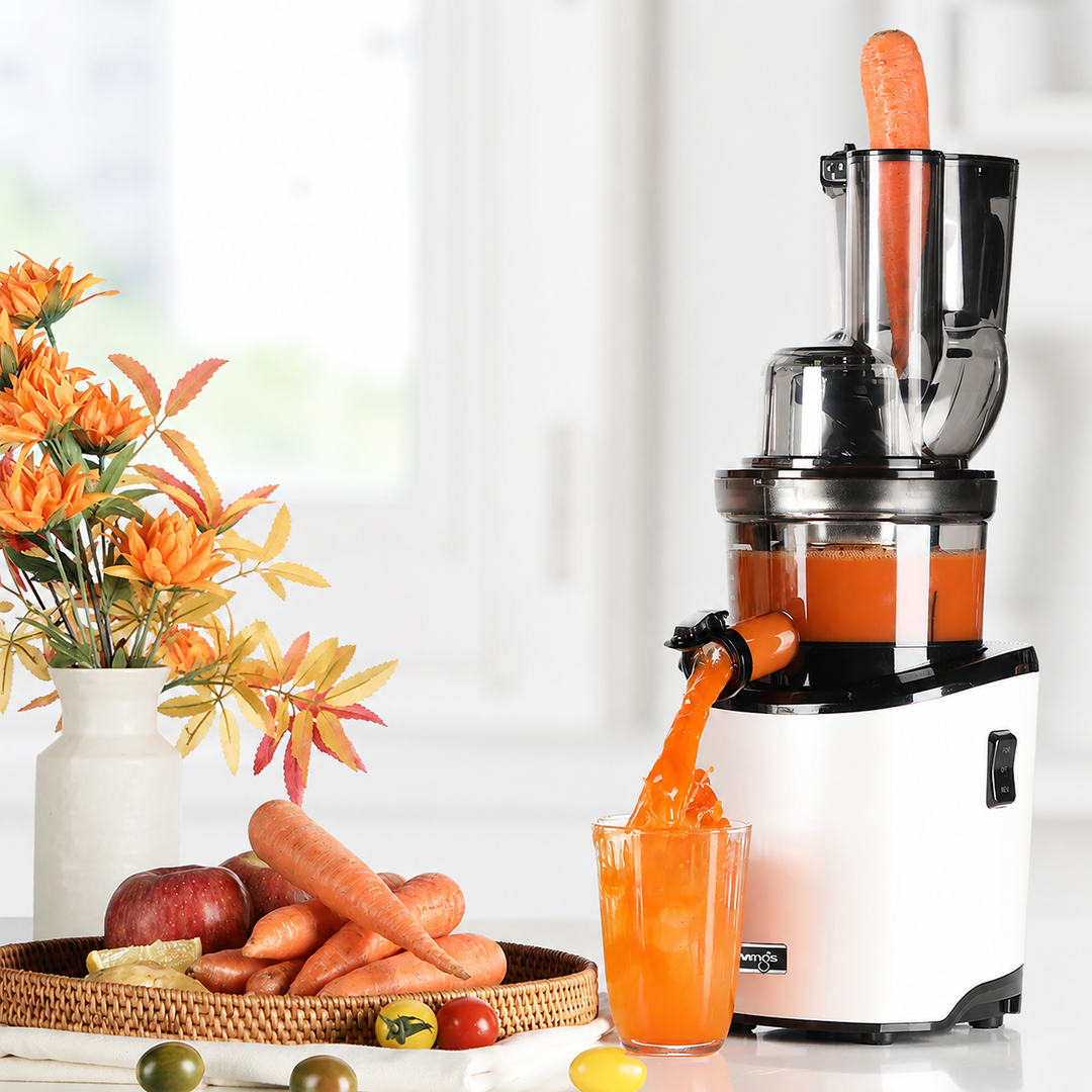 Eye-level lifestyle shot of a white Kuvings slow juicer processing a whole carrot in a bright kitchen, pouring fresh orange juice into a glass next to a wicker tray filled with carrots, apples, and tomatoes.