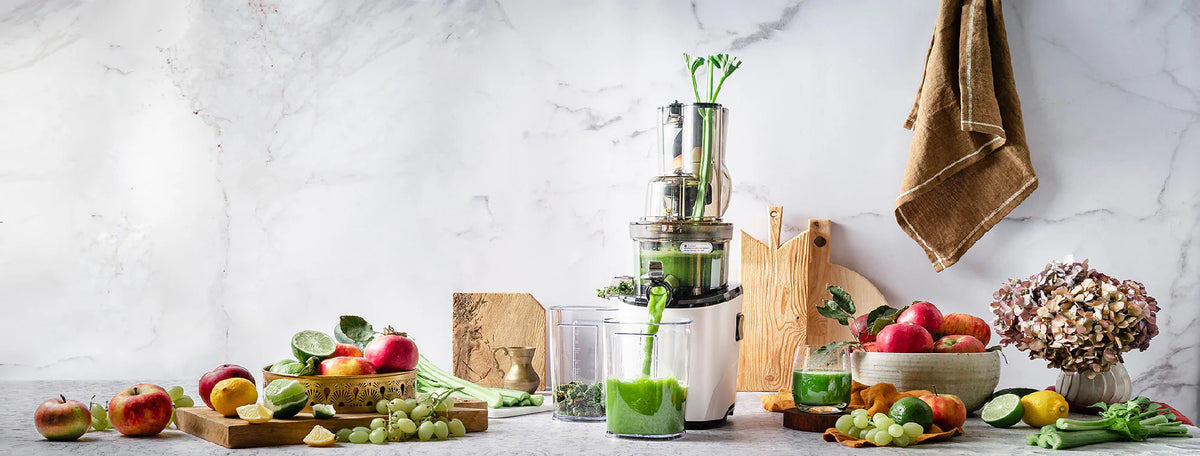 Wide panoramic lifestyle shot of a white Kuvings slow juicer extracting green celery juice on a marble kitchen counter, beautifully arranged with bowls of fresh apples, grapes, lemons, and lime slices.
